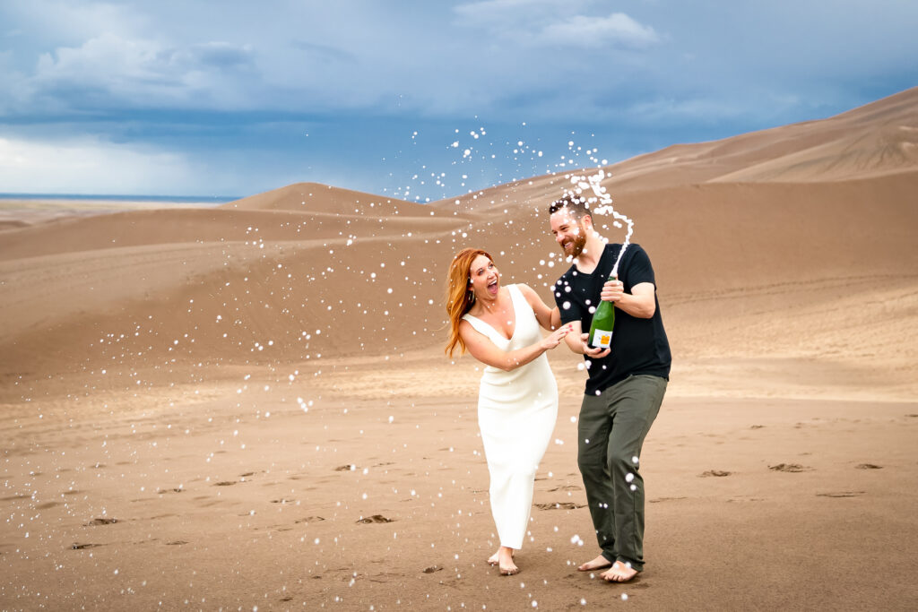 Engaged couple celebrates by popping a bottle of champagne in the sand dunes near the mountains, engagement inspiration, engagement photos, engagement photography, engagement photographer, engagement adventure, summer engagement session, Great Sand Dunes engagement inspiration, Great Sand Dunes engagement, Great Sand Dunes engagement photos, Great Sand Dunes engagement photography, Great Sand Dunes engagement photographer, Great Sand Dunes engagement adventure, Great Sand Dunes summer engagement session, Colorado engagement, Colorado engagement photos, Colorado engagement photography, Colorado engagement photographer, Colorado engagement adventure, Colorado summer engagement session, Mosca Colorado engagement, Mosca Colorado engagement photos, Mosca Colorado engagement photography, Mosca Colorado engagement photographer, Mosca Colorado engagement adventure, Mosca Colorado summer engagement session, Alamosa Colorado engagement, Alamosa Colorado engagement photos, Alamosa Colorado engagement photography, Alamosa Colorado engagement photographer, Alamosa Colorado engagement adventure, Alamosa Colorado summer engagement session, Sacred Whiteshell Mountain engagement photos