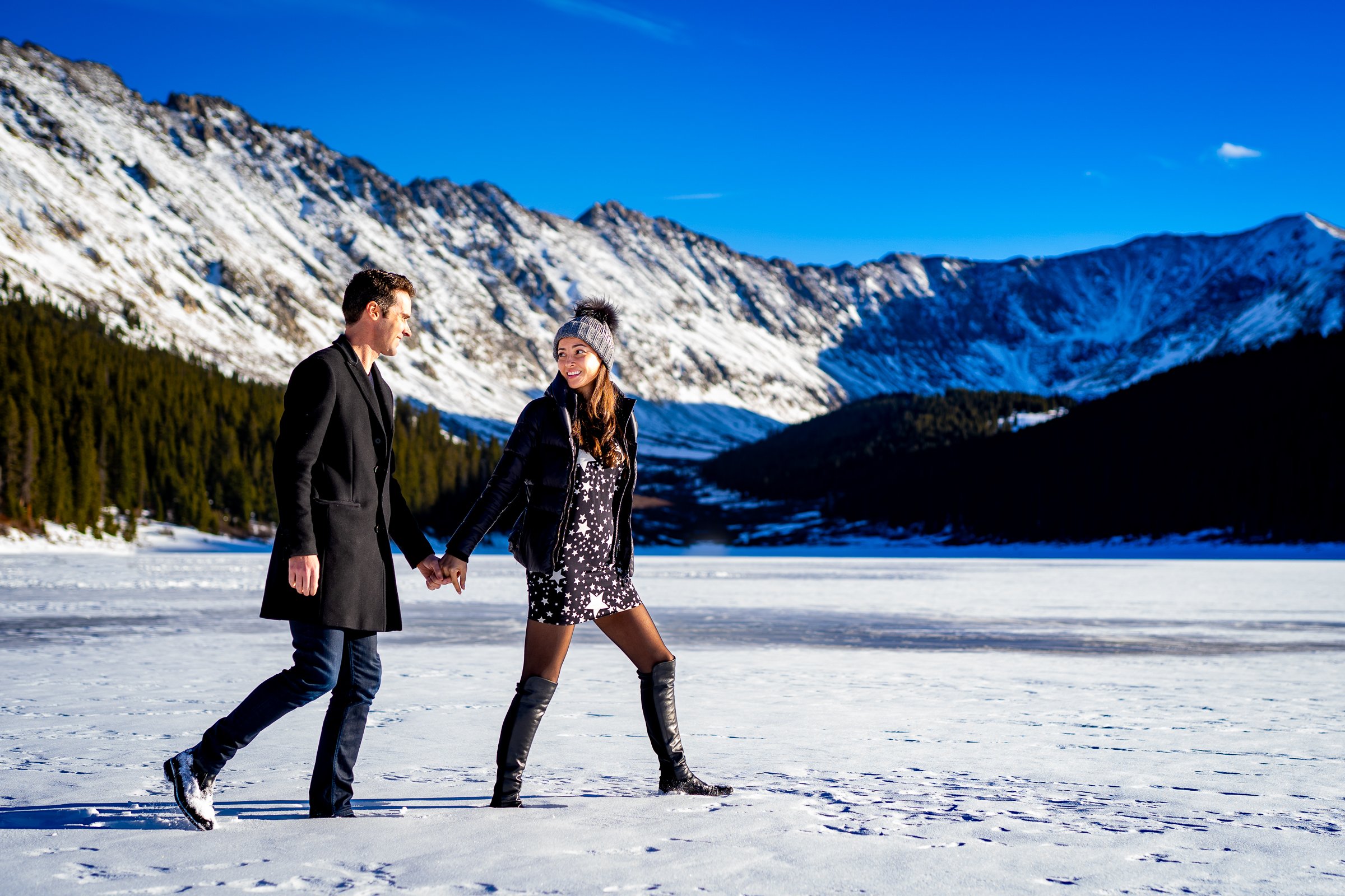 Engaged couple walks while holding hands on a frozen lake with snowcapped mountains in the background, Engagement Session, Engagement Photos, Engagement Photos Inspiration, Engagement Photography, Engagement Photographer, Winter Engagement Photos, Proposal Photos, Proposal Photographer, Proposal Photography, Winter Proposal, Mountain Proposal, Proposal Inspiration, Summit County engagement session, Summit County engagement photos, Summit County engagement photography, Summit County engagement photographer, Summit County engagement inspiration, Colorado engagement session, Colorado engagement photos, Colorado engagement photography, Colorado engagement photographer, Colorado engagement inspiration, Clinton Gulch Engagement