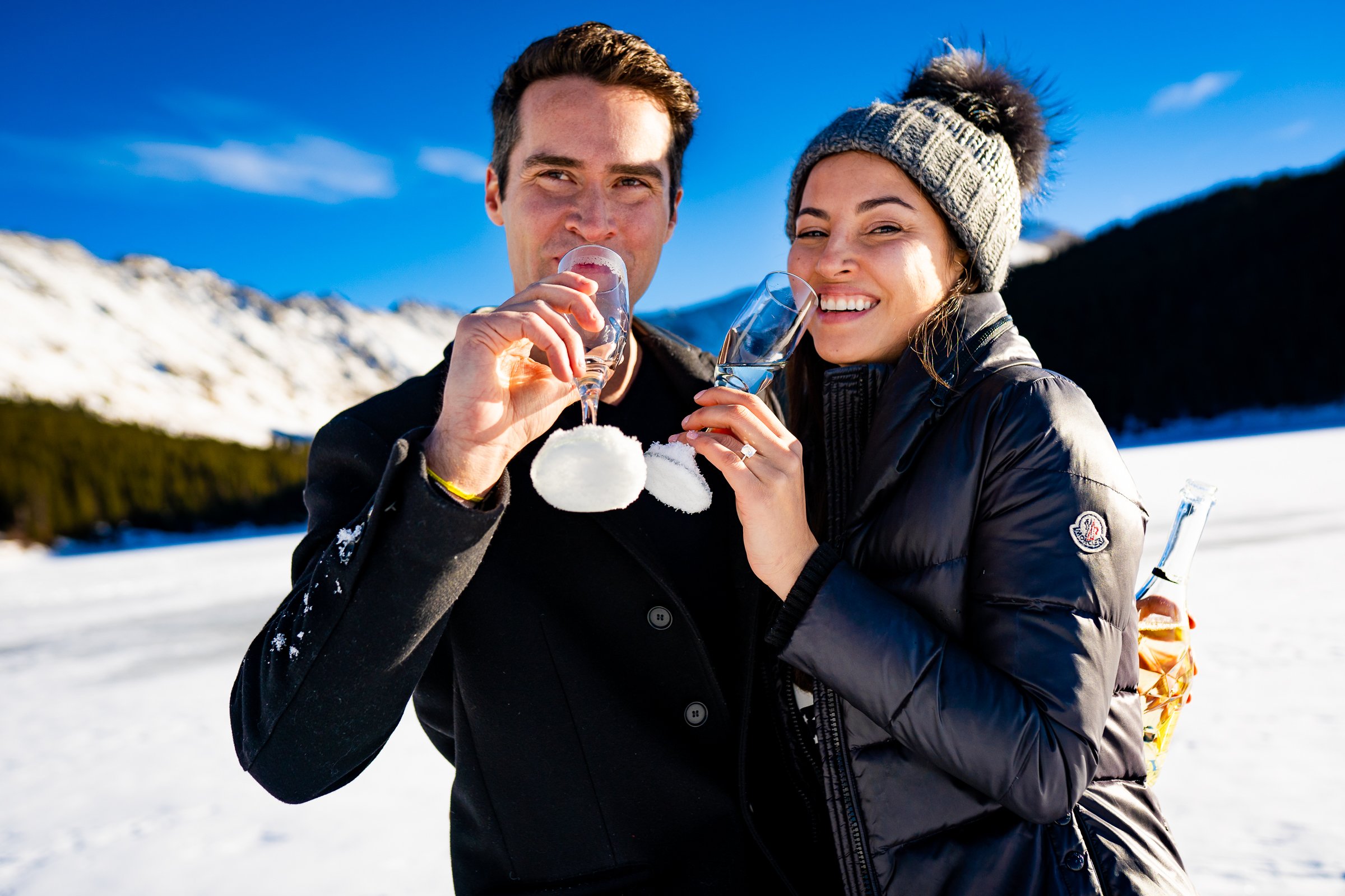 Engaged couple celebrates proposal with champagne on frozen lake with snowcapped mountains in the background, Engagement Session, Engagement Photos, Engagement Photos Inspiration, Engagement Photography, Engagement Photographer, Winter Engagement Photos, Proposal Photos, Proposal Photographer, Proposal Photography, Winter Proposal, Mountain Proposal, Proposal Inspiration, Summit County engagement session, Summit County engagement photos, Summit County engagement photography, Summit County engagement photographer, Summit County engagement inspiration, Colorado engagement session, Colorado engagement photos, Colorado engagement photography, Colorado engagement photographer, Colorado engagement inspiration, Clinton Gulch Engagement