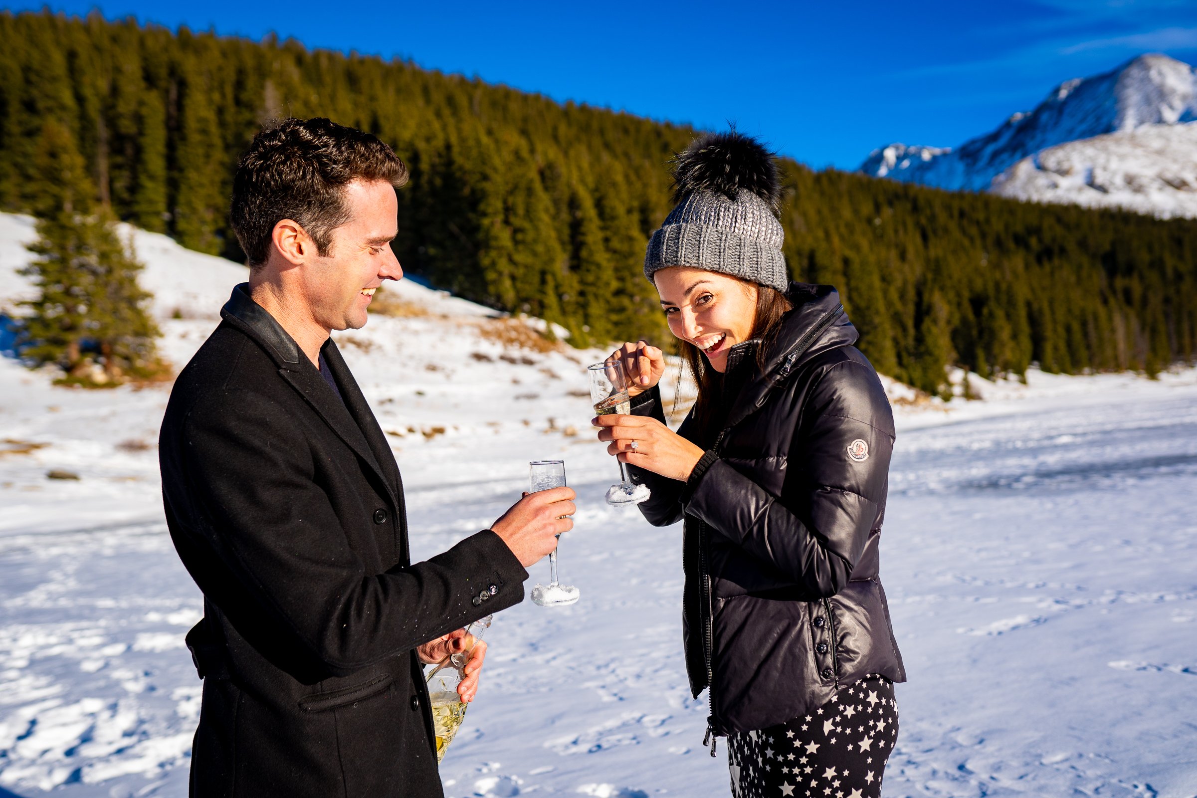 Engaged couple celebrates proposal with champagne on frozen lake with snowcapped mountains in the background, Engagement Session, Engagement Photos, Engagement Photos Inspiration, Engagement Photography, Engagement Photographer, Winter Engagement Photos, Proposal Photos, Proposal Photographer, Proposal Photography, Winter Proposal, Mountain Proposal, Proposal Inspiration, Summit County engagement session, Summit County engagement photos, Summit County engagement photography, Summit County engagement photographer, Summit County engagement inspiration, Colorado engagement session, Colorado engagement photos, Colorado engagement photography, Colorado engagement photographer, Colorado engagement inspiration, Clinton Gulch Engagement