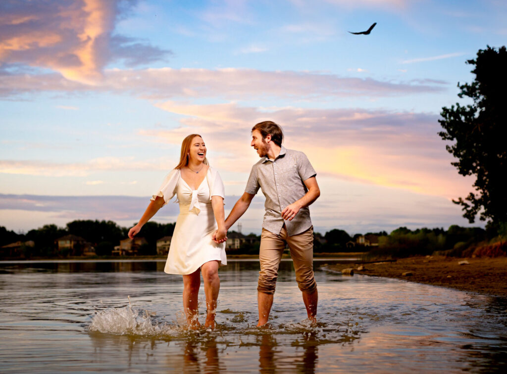 Newly engaged couple walks along the shore dipping their feet in the water at sunset.