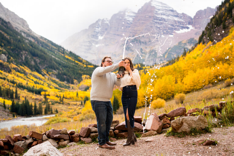 A newly engaged couple pops a bottle of champagne at Maroon Bells during peak fall foliage with gold aspens behind them