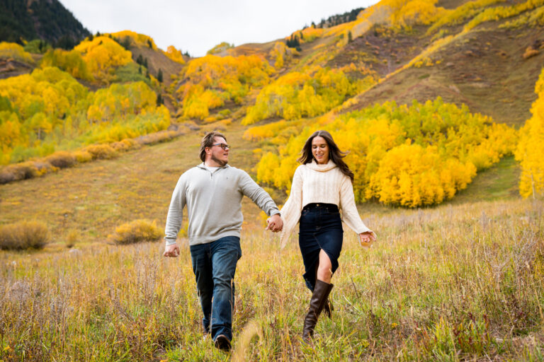 Couple walks through the meadow in front of a mountain glowing with fall colors and yellow aspens at Maroon Bells