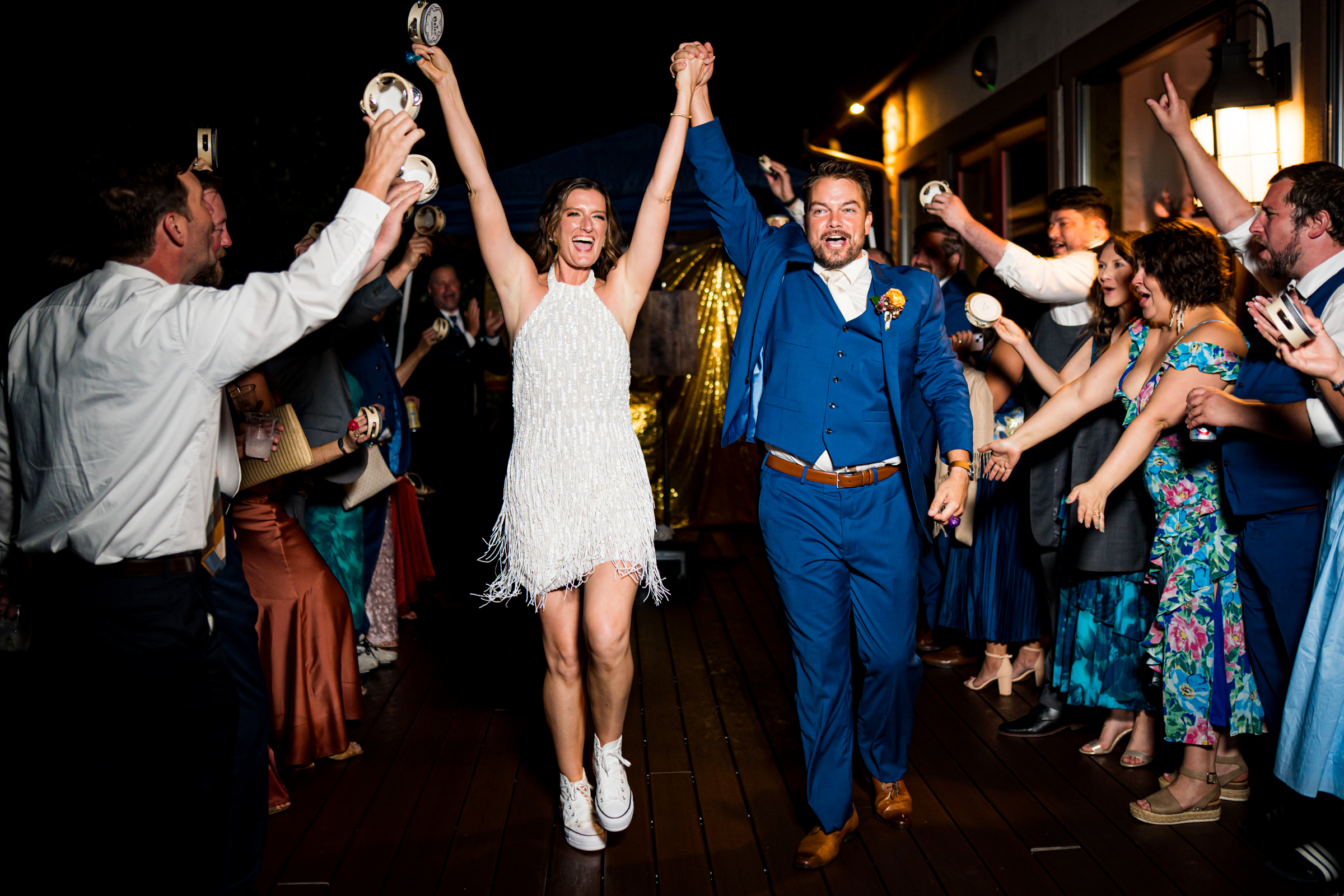 Bride and groom share a grand exit out of their reception hall, surrounded by guests who are shaking tambourines and cheering