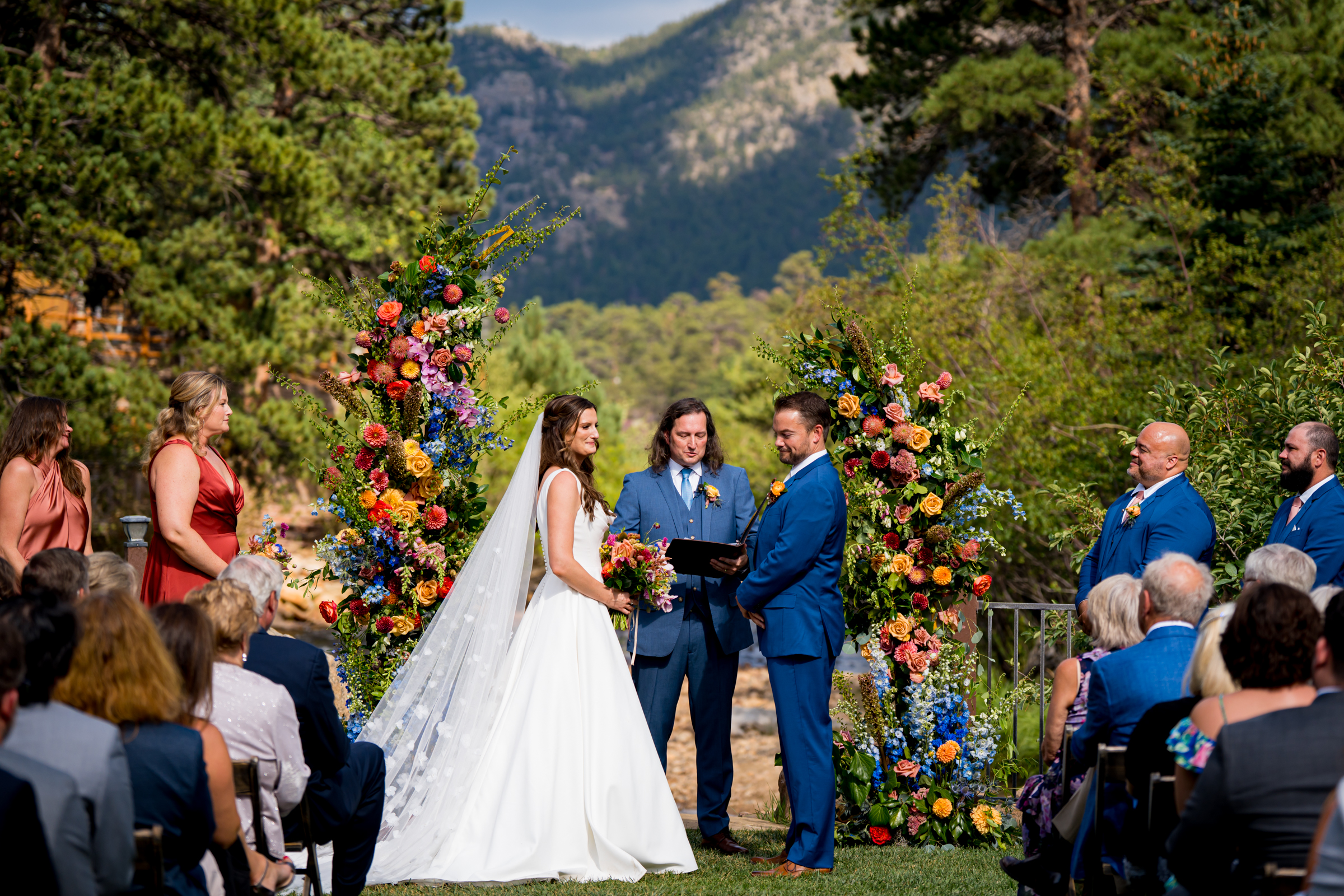 Bride and groom stand at the altar decorated in colorful summer flowers during there wedding ceremony, with the mountains and river as the backdrop
