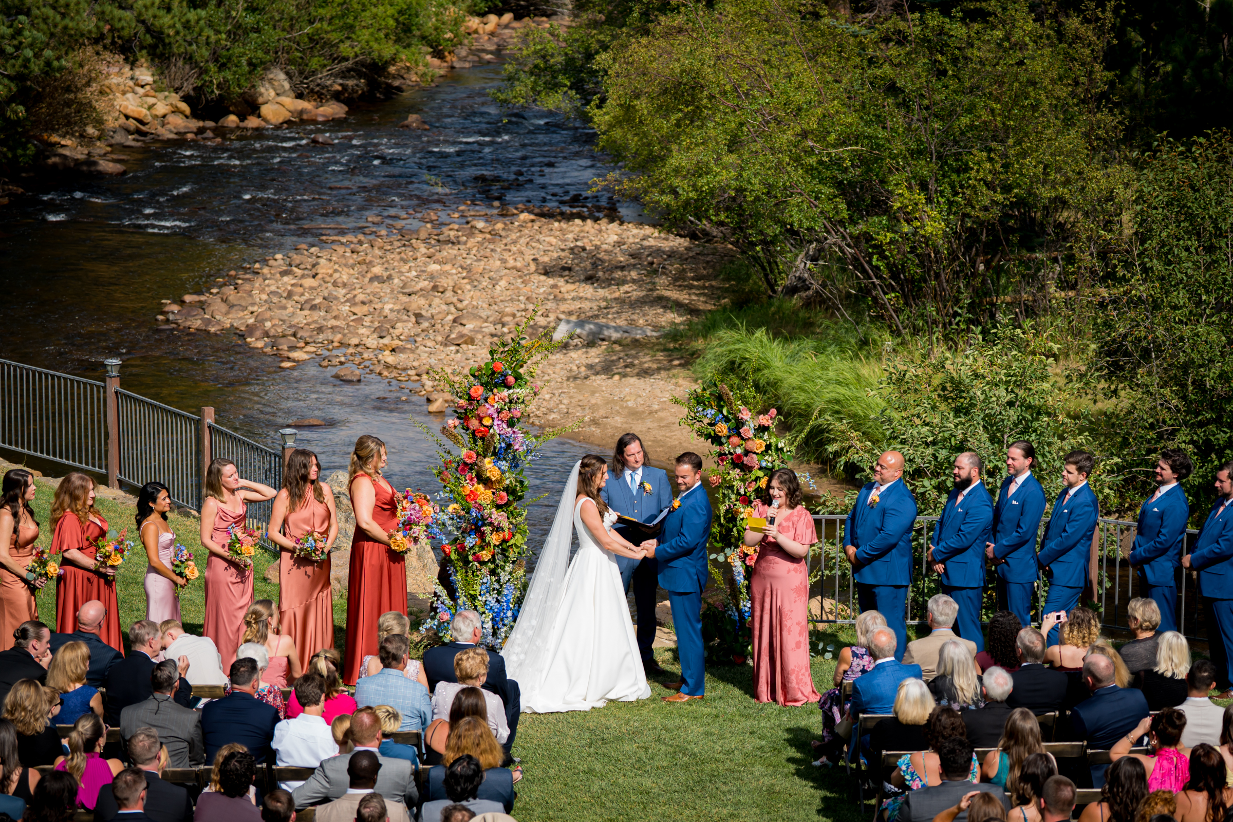 Bride and groom stand at the altar decorated in colorful summer flowers during there wedding ceremony, with the mountains and river as the backdrop