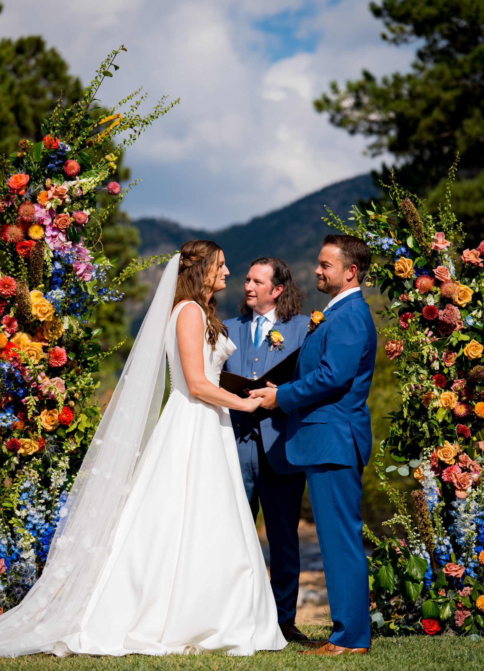 Bride and groom stand at the altar decorated in colorful summer flowers during there wedding ceremony, with the mountains and river as the backdrop