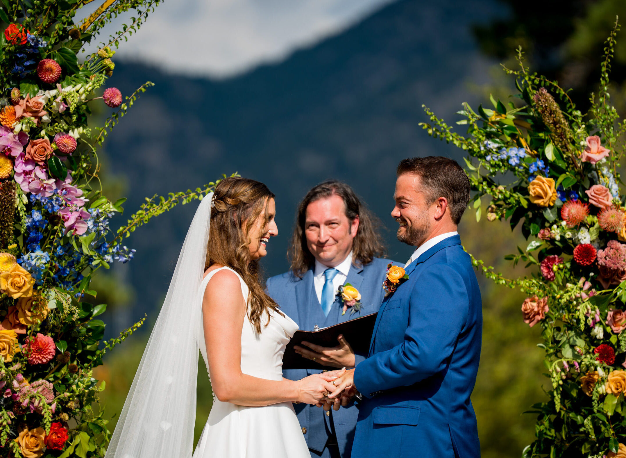 Bride and groom stand at the altar decorated in colorful summer flowers during there wedding ceremony, with the mountains and river as the backdrop