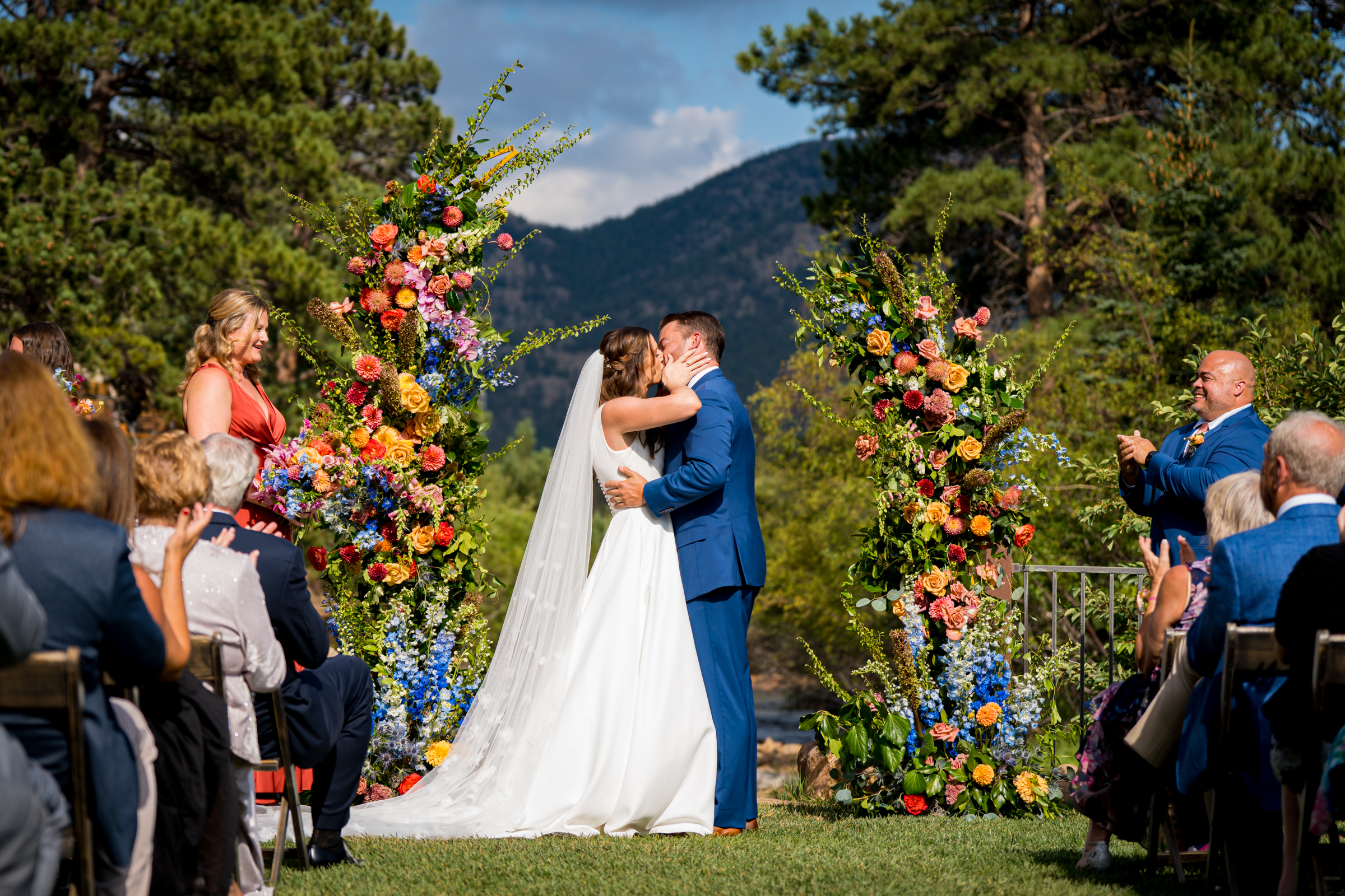 Bride and groom stand at the altar decorated in colorful summer flowers during there wedding ceremony, with the mountains and river as the backdrop