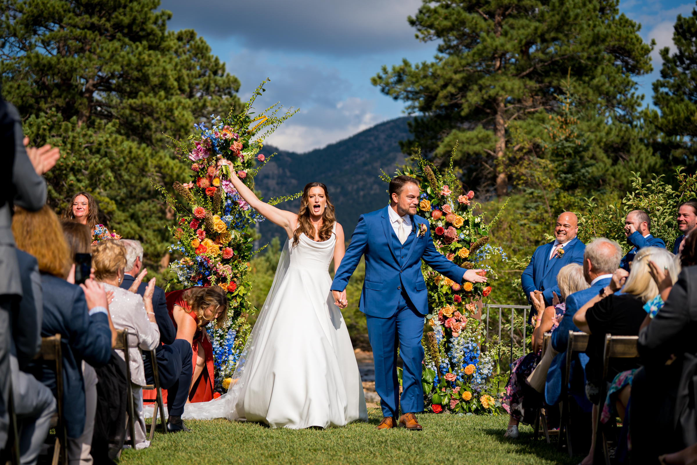 Bride and groom stand at the altar decorated in colorful summer flowers during there wedding ceremony, with the mountains and river as the backdrop